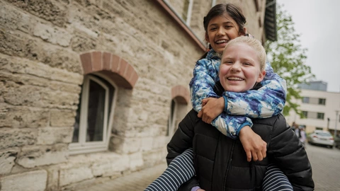 Eine Jugendliche, die lachend eine andere Jugendliche auf dem Rücken trägt.