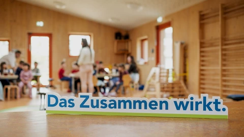 Slogan 'Das Zusammen wirkt' im Vordergrund. Hintergrund zeigt eine Gruppe von Kindern und Erwachsenen